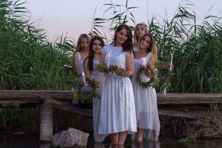 a group of young tangled girls in white dresses, with wreaths of flowers in their hands, are holding candles. On a natural background. A rustic style, a national holiday for young brides of Russia and Ukraine, Ivan Kupala, more truly the setting sun, young brides of twenty years old, standing by the water with wreaths and candles.の写真素材