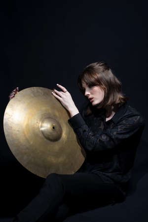 portrait of young beautiful smiling drummer girl holding drum big cymbal on black background.の写真素材