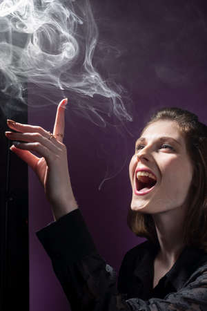 smiling young girl plays with her finger, touches the smoke of the vape. On a pink background in blackの写真素材