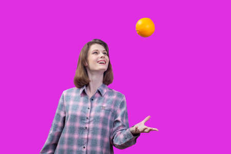 happy young girl tossing up an orange. Isolated on pink background Beautiful Caucasian little girl in a bright orange dress, throws a little ball.Isolated on white background.の写真素材