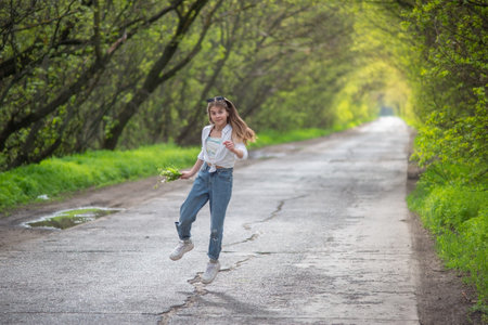 fashionable little girl standing walking on the road against the background of an arch of green branchy treesの写真素材