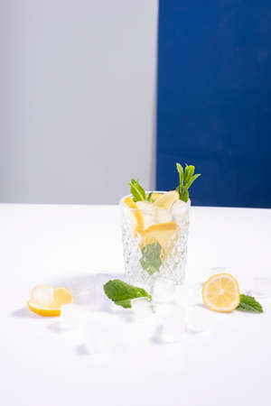creative summer cocktail with ice in a glass glass with lemon, mint and ice cubes on a white-blue background.の写真素材