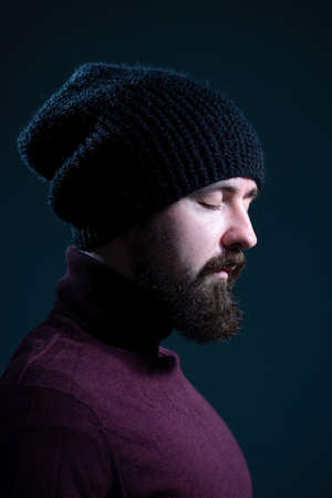 bearded guy with closed eyes in a knitted black hat and burgundy sweater. On a gray background.の写真素材