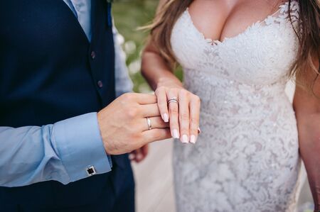 The groom in a suit and the bride in a white dress stand on the background of a green park and hold...