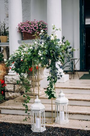 the arrangement of flowers at the wedding stands on a metal high stand, consists of many greens and white flowers stands on the steps of the stairs, next to it there are lanterns with candlesの写真素材