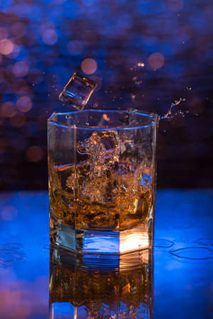 glass of whisky with dropped ice and splashes on a blue bokeh background.の写真素材