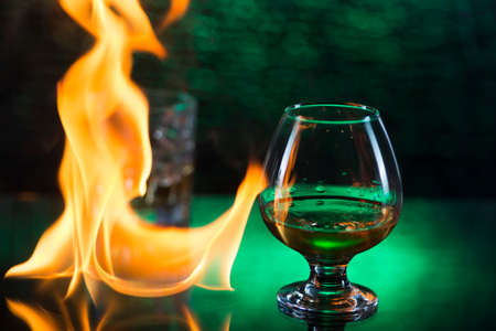 Glass o whisky with ice and wineglass of cognac and fire flames on green bokeh background.の写真素材
