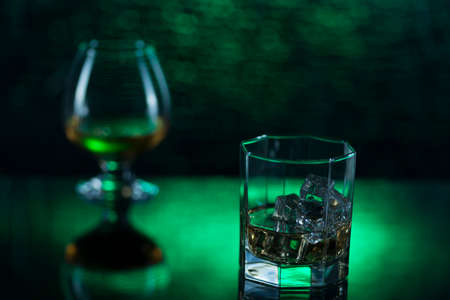 Glass o whisky with ice and wineglass of cognac on green bokeh background.の写真素材