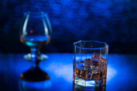 Glass o whisky with ice and wineglass of cognac on blue bokeh background.の写真素材