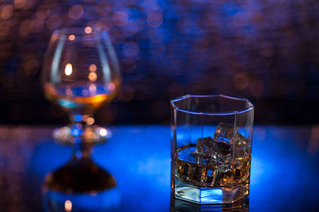 Glass o whisky with ice and wineglass of cognac on yellow-blue bokeh background.の写真素材