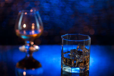 Glass o whisky with ice and wineglass of cognac on blue bokeh background.の写真素材