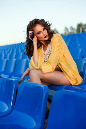 The beautiful girl with an elegant hairstyle sits at stadium against the background of blue seatsの写真素材