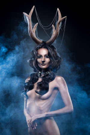 The naked beautiful girl with antlers on her head and a deer make-up in a blue fog. The breast is covered with hair. Deer horns entangled by web. Conceptual art photo. Halloween and Christmas.の写真素材