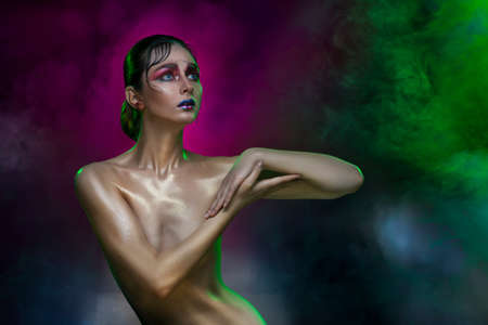 The beautiful naked slim girl covers a breast with hands in multi-colored theatrical smoke. Clean healthy wet shining skin. Vanguard make-up and hairstyle. Copy space.の写真素材