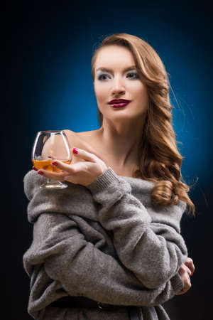 Beautiful elegant girl wearing a gray sweater holds in hands a glass of cognac. Copy space. Glamourous hairstyle. Red lips. Evening make-up. Healthy clean skin. Blue-black gradient backgroundの写真素材