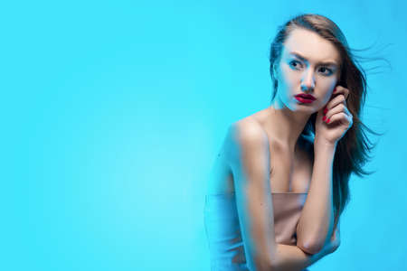 The beautiful blonde red lips girl touches by fingers face and a neck and scared looks sideways.Blue background. Healthy clean leather. Manicure - red nails. Copy space.の写真素材
