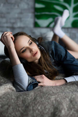 The beautiful girl laying on a bed wearing underwear and a jeans jacket listens to music via earphones. Copy space. Advertising and Commercial Design, Fashion. Healthy clean skin.の写真素材