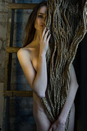 Beautiful naked girl, shyly covering her nakedness with a bunch of ropes in the loft-style room. Copy space. Advertising, fashion and commercial Design, Fashion. Healthy clean skin.の写真素材