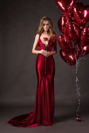 Beautiful woman wearing a deep neckline red satin dress holding red rose in her hand posing with red balloons in the shape of a heart.の写真素材