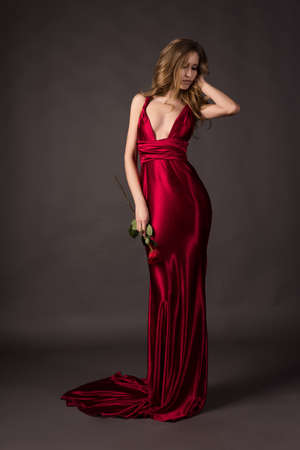Beautiful woman wearing a deep neckline red satin dress holding red rose in her hand on a grey background.の写真素材