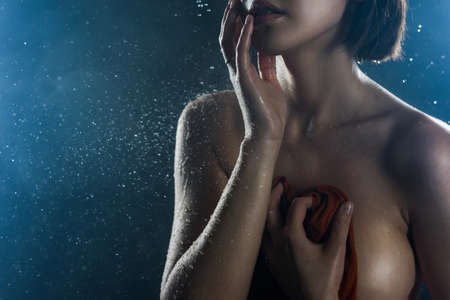 Beautiful big breasted girl covers herself with a wet shirt on a dark background. Falling rain drops and artistic scenic smoke. Advertising, commercial design. Copy space. Mixed Asian-Caucasian race.の写真素材