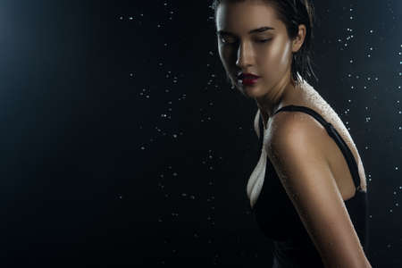Beautiful wet big breasted girl wearing black swimsuit on a dark background. Falling rain drops and...