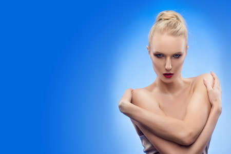 The beautiful blonde big breasted girl embraces herself. Blue background. Evening makeup. Healthy...