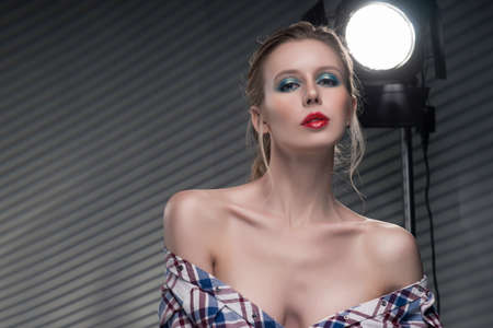 Beautiful slender blonde big breast girl without a bra wearing unbuttoned checkered shirt and jeans on a striped gray background near the studio lamp. Copy space. Advertising, fashion, and commercialの写真素材