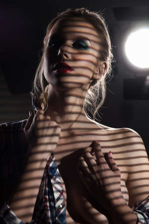 A beautiful slender blonde big breasts girl without a bra wearing an unbuttoned checkered shirt and jeans covered with shadows from the blinds near the studio lamp. Copy space. Grey background.の写真素材
