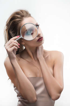 Beautiful naked shoulders girl looking through a magnifying glass and increases with it her beautiful eye.  Healthy clean skin. Isolated on white background. Cosmetology, spa, skin care. Copy space.の写真素材