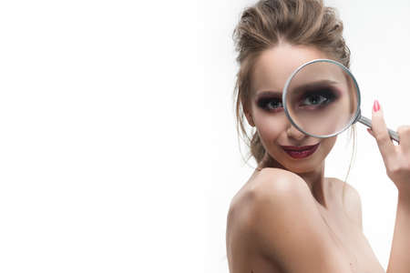 Beautiful naked shoulders girl looking through a magnifying glass and increases with it her beautiful eye.  Healthy clean skin. Isolated on white background. Cosmetology, spa, skin care. Copy space.の写真素材