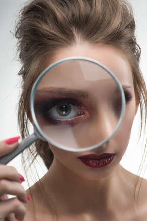 Beautiful naked shoulders girl looking through a magnifying glass and increases with it her beautiful eye.  Healthy clean skin. Isolated on white background. Cosmetology, spa, skin care. Copy space.の写真素材