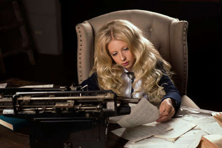Beautiful long haired curly blonde schoolgirl tiredly learns homework in a classic vintage interior room, sitting at a table at a typewriter among books and sheets of text. School, education.の写真素材