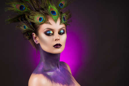 Beautiful big breast girl wearing peacock feathers in her hair and artistic violet shiny body art on her neck, vanguard makeup. Copy space. Violet background. Conceptual, advertising and commercial.の写真素材