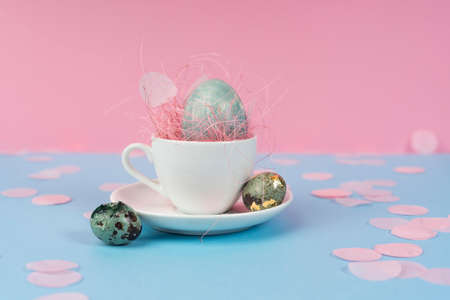 Conceptual art photo of a blue chicken and quail eggs in a white coffee cup on a saucer, decorated with a pink sisal and confetti on a blue and pink background. Copy space. Advertising design.の写真素材