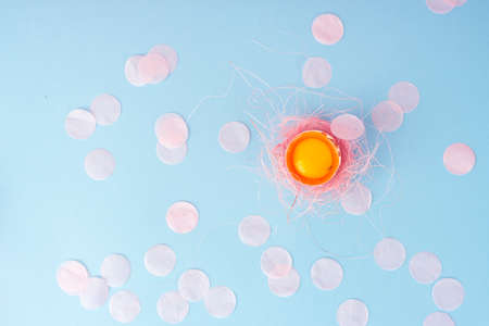 Yolk of broken egg in eggshell decorated with pink sisal nest and pink confetti on the minimalist  blue background. Copy space. Advertising, commercial design.の写真素材