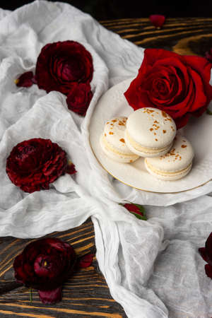 Three macaroons pastry lying on a plate surrounded by rose blossoms and rose petals on a wooden table covered with a crumpled white cloth. Still life.  Advertising, commercial design. Copy space.の写真素材
