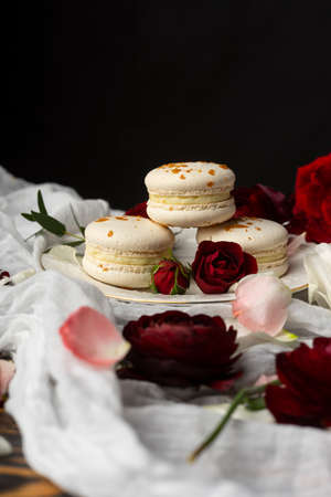 Three macaroons pastry lying on a plate surrounded by rose blossoms and rose petals on a wooden table covered with a crumpled white cloth. Still life.  Advertising, commercial design. Copy space.の写真素材
