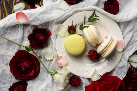 Three macaroons pastry lying on a plate surrounded by rose blossoms and rose petals on a wooden...