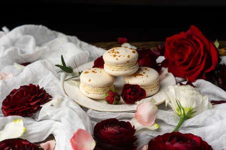 Three macaroons pastry lying on a plate surrounded by rose blossoms and rose petals on a wooden table covered with a crumpled white cloth. Still life.  Advertising, commercial design. Copy space.の写真素材