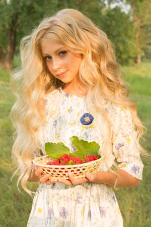 Beautiful charming long curly blonde hair teenage girl wearing a long light dress outdoors on a picnic holds a plate full of raspberries in her hands. Copy space. Lifestyle.の写真素材