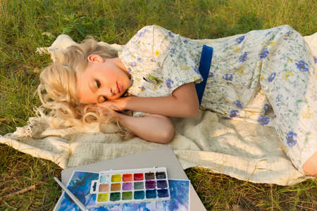 Tired beautiful charming barefoot blonde teenage painter girl wearing a long light dress outdoors sleeps on a blanket on the grass next to the brush, watercolor and a sketch. Copy space. Lifestyle.の写真素材