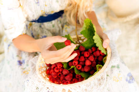 The hands of a blond teenage girl hold a full little basket of raspberry berries lying on large...