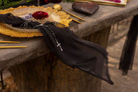 A red rosebud lay on a golden plate next to a black veil with a necklace belt with a crossed wind, a...