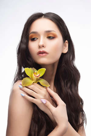 A beautiful brunette nude shoulders girl sensually holds a flower of an orchid in her hands. Nude make-up. Healthy smooth skin. Isolated on white background. Copy space. Commercial and advertising.の写真素材