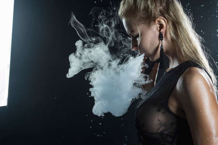Beautiful shapely athletic big breasted  blonde girl with wet oiled body wearing black translucent sexy bodysuit vaping vape in scenic smoke under falling water drops of rain on black. Copy spaceの写真素材
