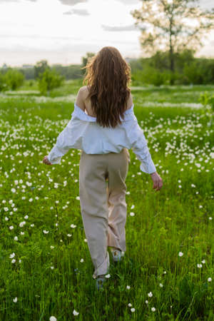 A beautiful slim girl wearing a fashionable white shirt, and beige trousers goes away into the sunset in the green meadow among the wild flowers. Copy space. Commercial and advertising design.の写真素材