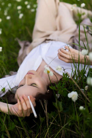 Beautiful stylish young girl wearing a trendy white shirt, beige bloomers, and a pareo lies relaxing on the green grass among the wild flowers and smoking cigarette. Advertisement design. Copy space.の写真素材