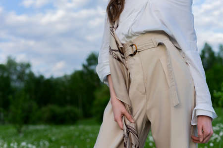 Beautiful stylish girl wearing a trendy white shirt, beige trousers and a pareo poses outdoors against a background of meadow full of wild flowers and blue cloudy sky. Advertising design. Copy space.の写真素材
