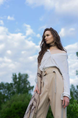 Beautiful stylish girl wearing a trendy white shirt, beige trousers and a pareo poses outdoors against a background of meadow full of wild flowers and blue cloudy sky. Advertising design. Copy space.の写真素材